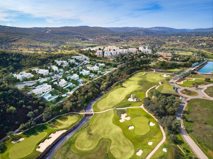 An aerial view of a golf resort with fairways, villas, winding paths, and a reflective water feature, set against rolling hills and a bright blue sky