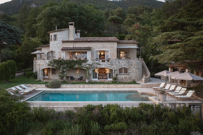 A stone villa with warm lights, arched windows, a terracotta roof, and a large pool, surrounded by trees and mountains