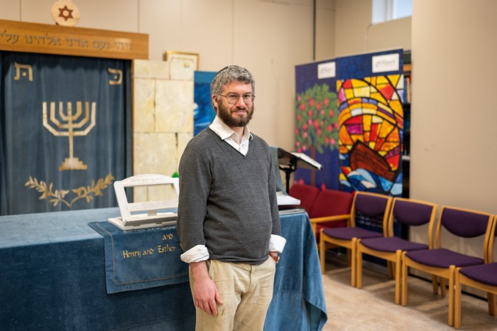 A man standing inside a synagogue near a Torah ark, with chairs and stained-glass artwork in the background