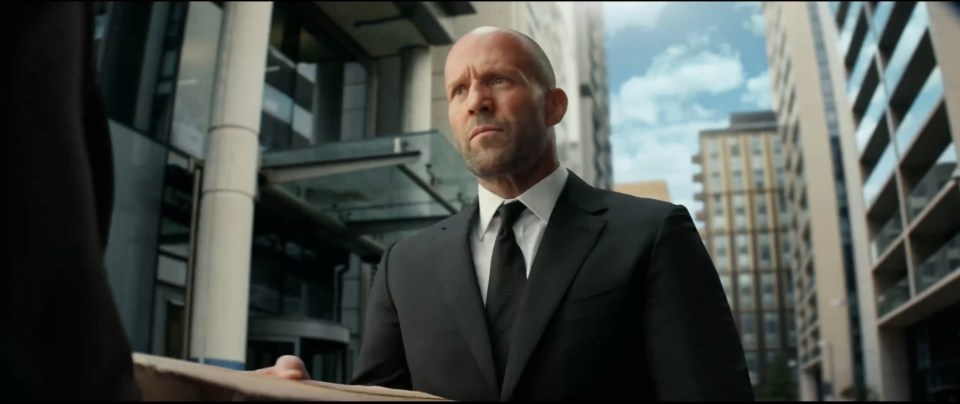 Jason Statham in a suit, handing over a package.