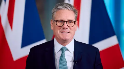 Getty Images  Britain's Prime Minister Keir Starmer speaks at a press conference, during his visit to the European Commission headquarters on October 2, 2024 in Brussels, Belgium. Behind Sir Keir are two flags of the UK in the distance. He is wearing a navy suit, black spectacles and a blue tie.