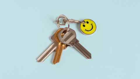 A set of three metallic keys, one gold and two silver, is attached to a keyring with a yellow smiley face keychain, placed against a light blue background