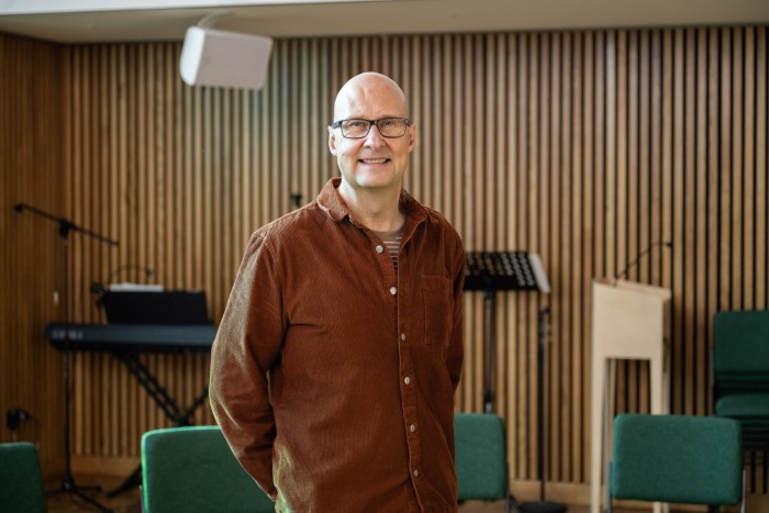 A smiling man stands in a modern room with wooden paneling, green chairs, and musical equipment in the background