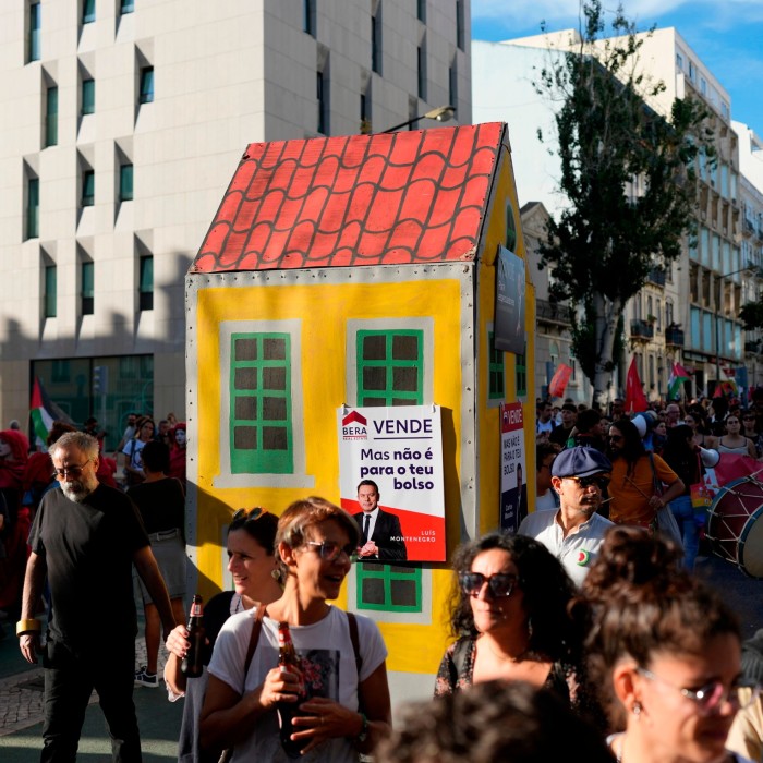 A lively protest with people marching around a small, yellow, cartoonish house prop displaying a political sign about housing prices