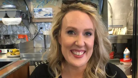 Catherine Thom Catherine Thom with blonde wavy shoulder length hair and blue eyes smiling at the camera wearing a black top in a cafe with the kitchen seen behind her