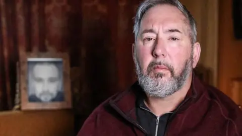 Mike Haines stares straight at the camera in a close-up. He is wearing a maroon jacket and a black zip-up top underneath. He has short grey hair and a beard. There is a framed photo in the background.