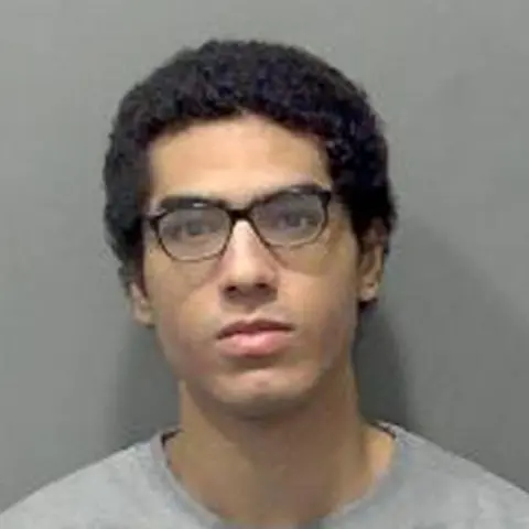 PA Nicholas Prosper looks straight ahead in a police custody photo. He is wearing a grey T-shirt and has dark short hair and glasses