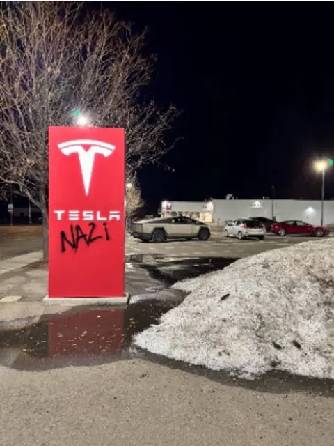 US Department of Justice A Tesla dealership with the word "Nazi" spray painted on the sign. Cars and snow banks are visible in the photo.