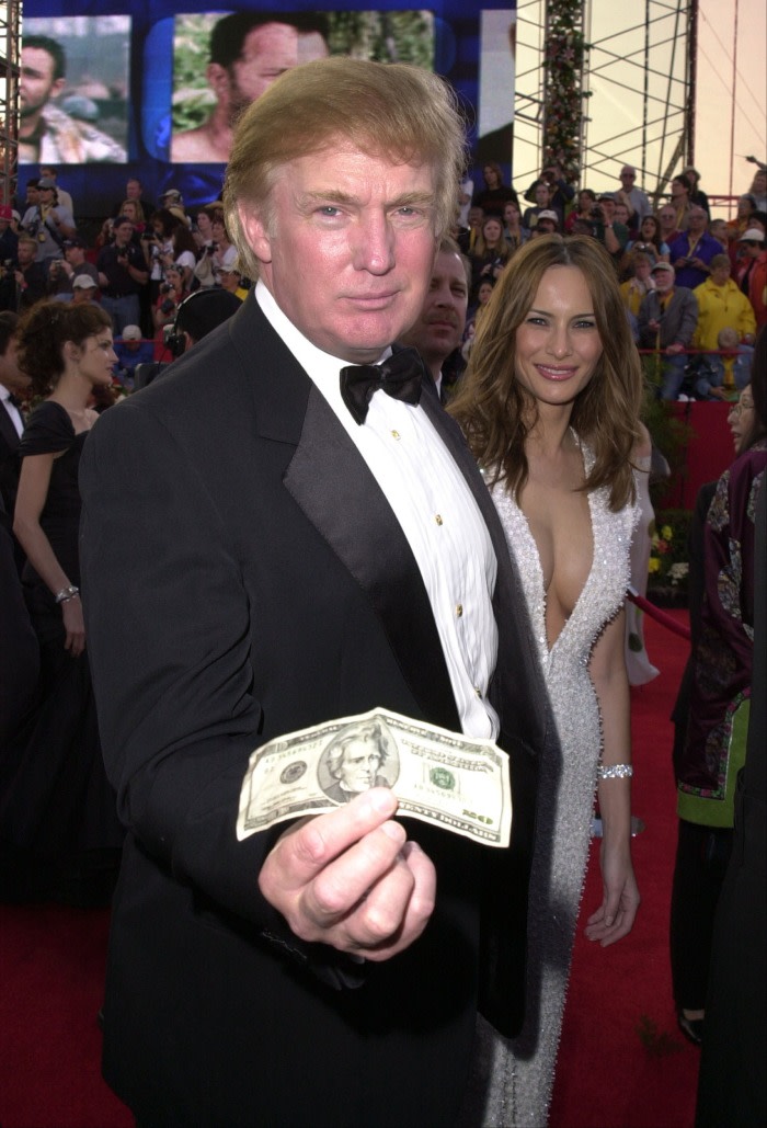 A man in a tuxedo holds a dollar bill towards the camera. Behind him stands a smiling young woman in a low-cut evening dress