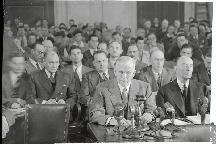 In a room full of men in 1940s-style suits, a man sits at a desk, with a microphone in front of him