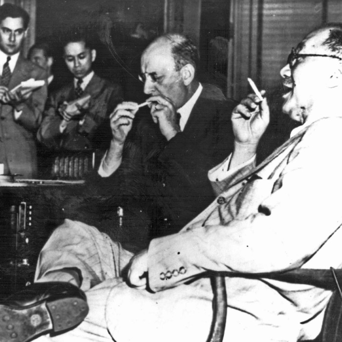 Two men sit smoking, the man in the foreground leaning back in his chair