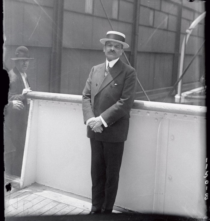 A man in a double-breasted suit and boater leans against a white wall on the deck of a ship on a sunny day 