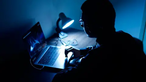 Getty Images A man whose face is in shadow sits in front of a laptop