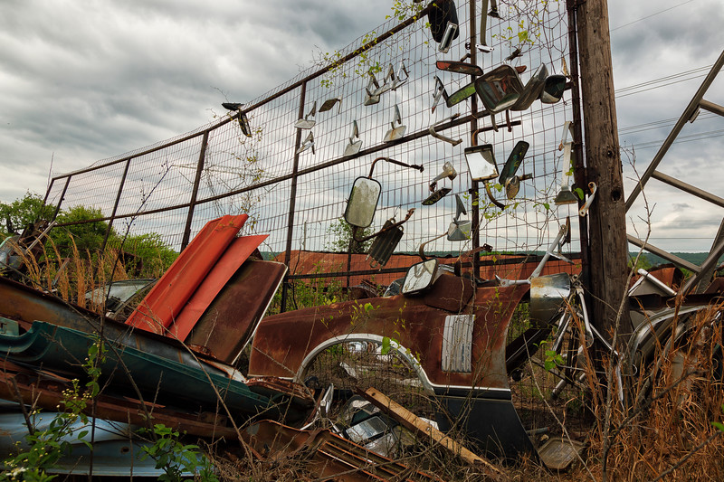 Abandoned graveyard of luxury cars with car parts attached to a fence.