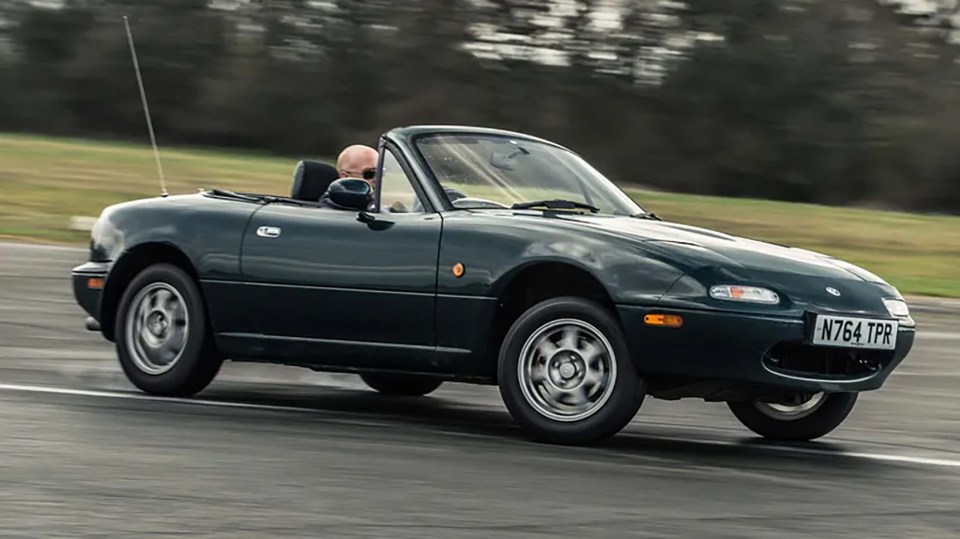 A dark green Mazda MX-5 (NA) convertible driving on a track.