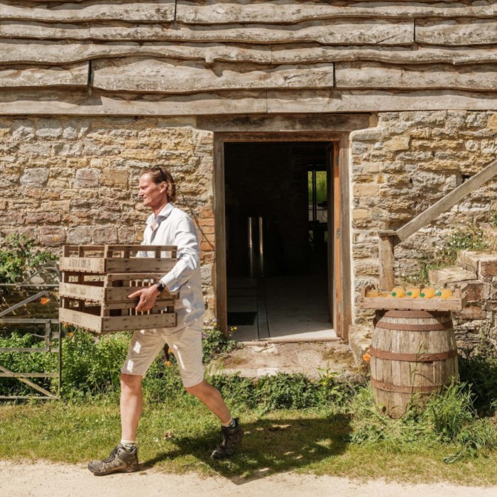 Man in light clothing carrying wooden crates outside a rustic stone building with timber cladding