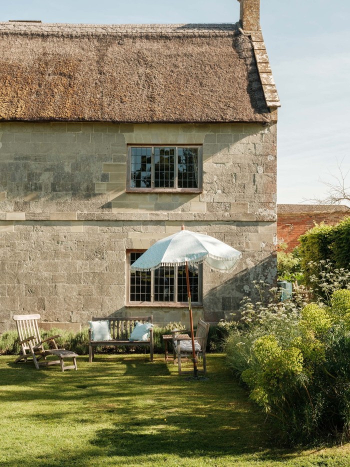 Stone cottage with thatched roof, shaded garden seating and a parasol set on a neatly mown lawn