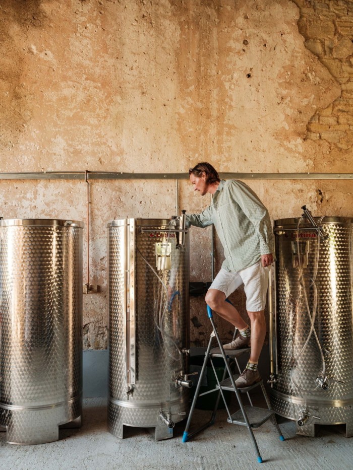 Man in light clothing standing on a step ladder inspecting stainless steel fermentation tanks in a rustic room