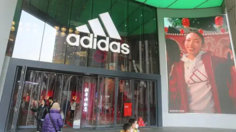 Getty Images Adidas shopfront. People are walking through the front doors of a big glass shopfront with the adidas logo above it. The logo is three left-facing rectangles at 45 degrees increasing in size from left to right. On the right of the shop is a massive banner of a model happily wearing an Adidas hooded top. She is smiling broadly.
