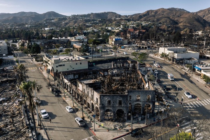 Charred remains of buildings stand in a scorched city block, surrounded by intact streets and distant hills