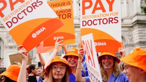 PA Media Junior doctors protest in August 2023, wearing blue scrubs and orange hats hold placards reading: "Pay restoration for doctors".