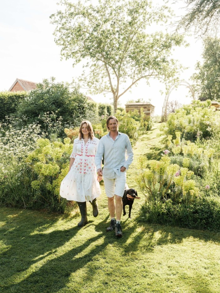Man and woman walking hand in hand with a black dog through a sunny garden path lined with wildflowers